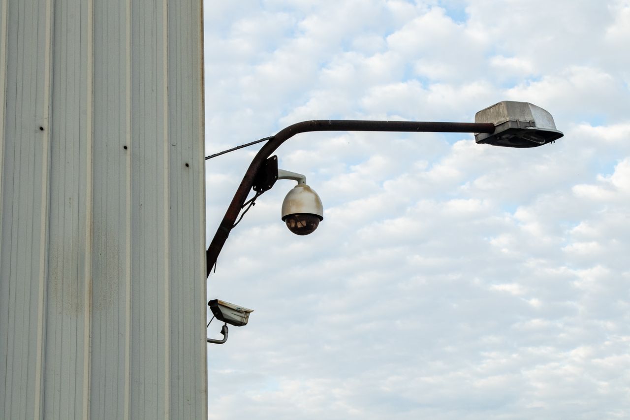 Street lamp and CCTV camera mounted on a building against a cloudy sky.